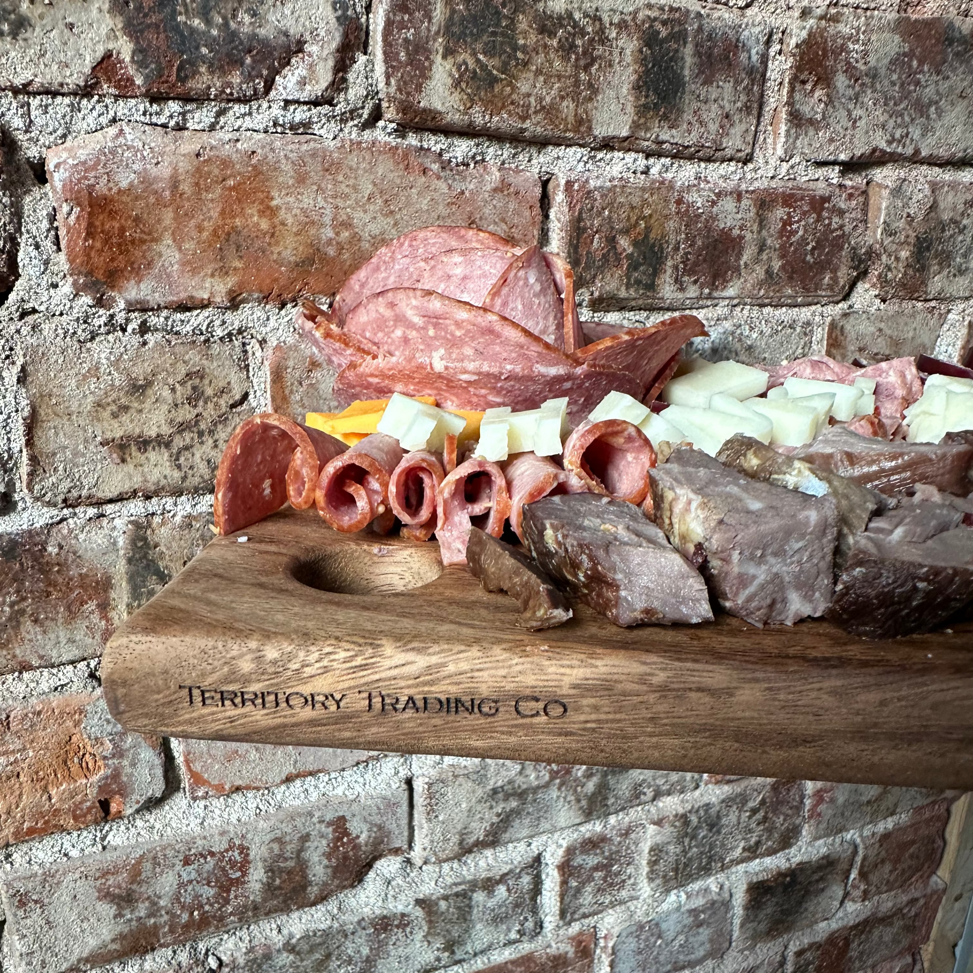 Wooden charcuterie board with meats and cheeses against a brick wall.
